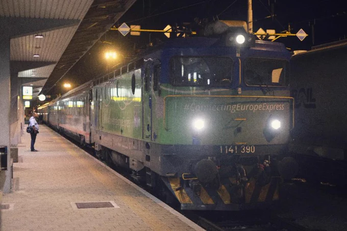 The HŽ loco in front of the train in blue and green livery with headlights switched on waiting for departure. The steward is waiting on the platform.