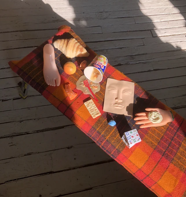  A photograph of objects on an orange, red, and blue plaid bench outside. The objects include a cup of noodles, crackers, a croissant, a hand figurine, and more. 
