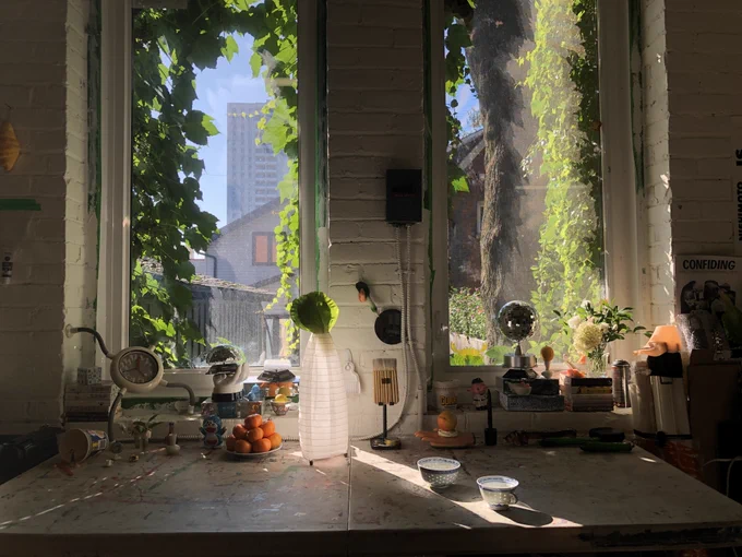 A photograph of objects on a table placed beneath two large windows. The objects include oranges, bowls, vases, plants, a clock, and more. 
