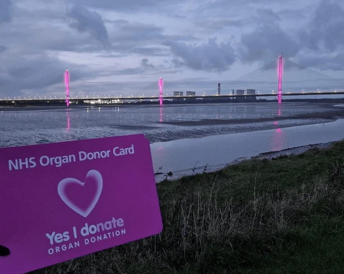A photo of Mersey Bridge lit up pink from a distance. In the foreground is a large pink NHS Organ Donor Card being held up.