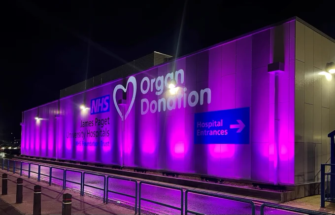 A photo of the outside of a building belonging to James Paget University Hospitals NHS Foundation Trust with pink lights and a projection of the Organ Donation logo on.