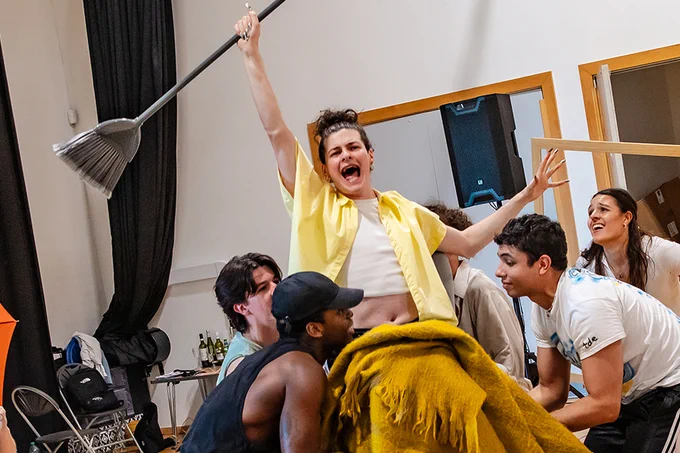 A photo showing a group of performers in rehearsals with a performer holding a broom and shouting