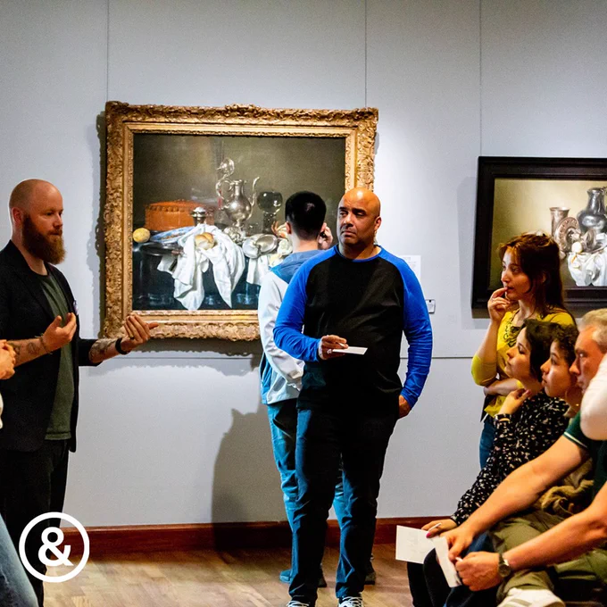 Foto: een rondleider vertelt iets aan een groep luisteraars. Ze staan in een museum, voor klassieke kunstwerken.