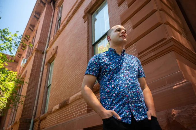  Portrait of Adam Powell taken from below. Powell is standing in front of a red brick building wearing a blue patterned shirt and black pants. His hands are in his pockets as he looks off into the distance. Photo by Leila Saidane. 