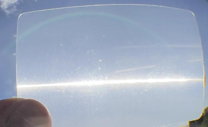 Scheckkartengroße, längsstrukturierte Folie gegen die Sonne gehalten. Die Sonne erscheint durch die Folie als Lichtstreifen.