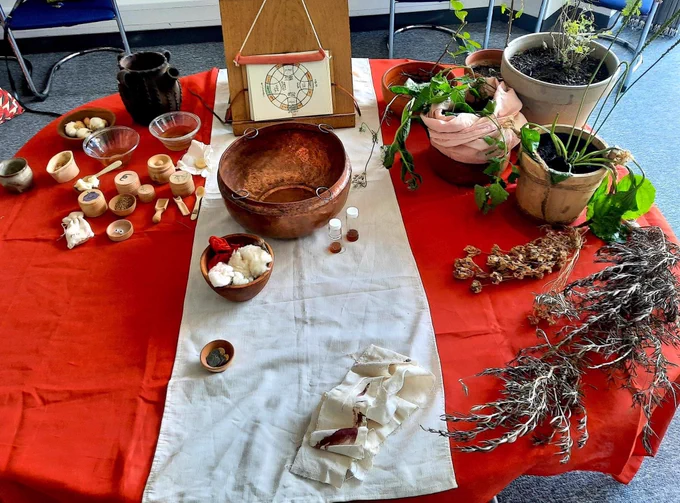 A table covered with live and dried plants, a book illustrating the elements and humours, and various bowls and pots of substances.