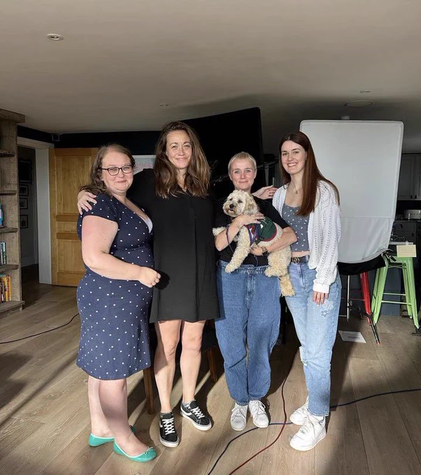A photo of four people standing next to each other and smiling. One is holding a small light furry dog with a green harness on. In the background is filming equipment.