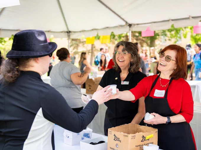  UNM President Garnett Stokes, at far right in red with a black apron, hands a cup of ice cream to a Lobo student in a long-sleeve black and white shirt and wearing a fashionable black fedora. UNM EVP Teresa Costantinidis, in black, looks on at center.