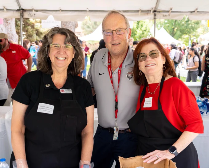 UNM President Garnett Stokes at far right, in red shirt and black apron, wearing New Mexico jewelry, with her husband Jeff at center, in gray Lobo shirt, and UNM EVP for Finance & Administration Teresa Costantinidis on the left in a short-sleeved black Lobo with a UNM pin on the lapel.
