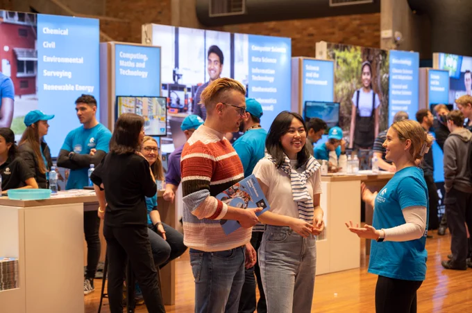 Three people, one man and two women, are facing each other having a conversation. They are inside, and behind them is a crowd of people standing at white desks and blue screens.