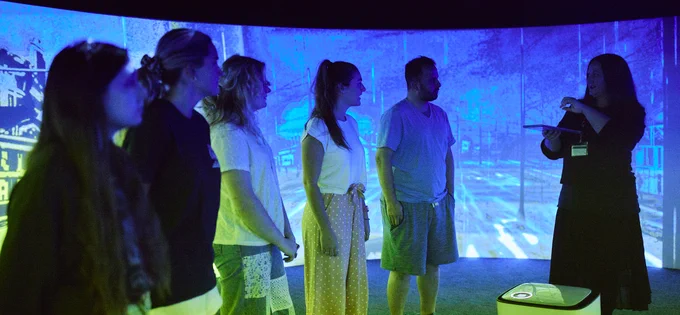 Five people are facing a woman who is speaking to them and they are standing inside a dark room that has large screens projecting blue and green imagery on the walls.