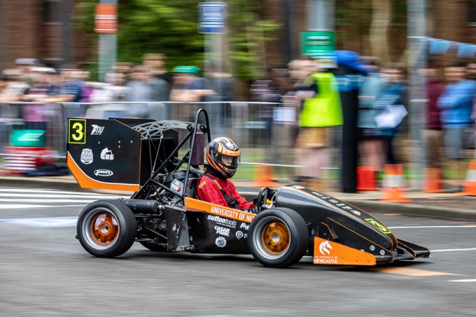 A person with a black helmet and red shirt on is sitting in a black and orange race car. The race car is moving on the black road, and there is a crowd of people watching from behind.