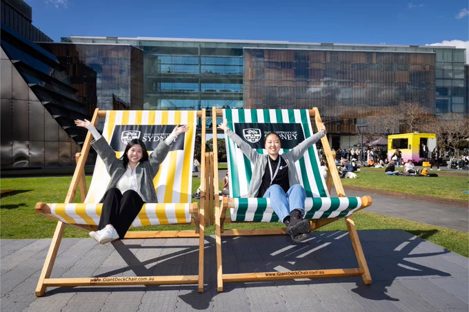 Two students sit on oversized deck chairs one on left is yellow and right is blue. They are throwing their hands up and smiling