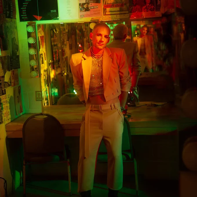 Image of ted in the buddies dressing room, photographed in green and red lighting. He is wearing a cropped white blazer with a sequin shirt underneath and a pearl necklace, and white pants with black boots. He is leaning against the back of a chair, with a mirror behind him.