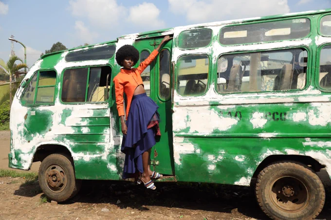 This image shows a model wearing an afro hairdo hanging from the closed door of a green weathered minibus. The model wears an orange cropped shirt and a navy-blue multi-layered high-low skirt with accents of  a maroon and orange checkered fabric.