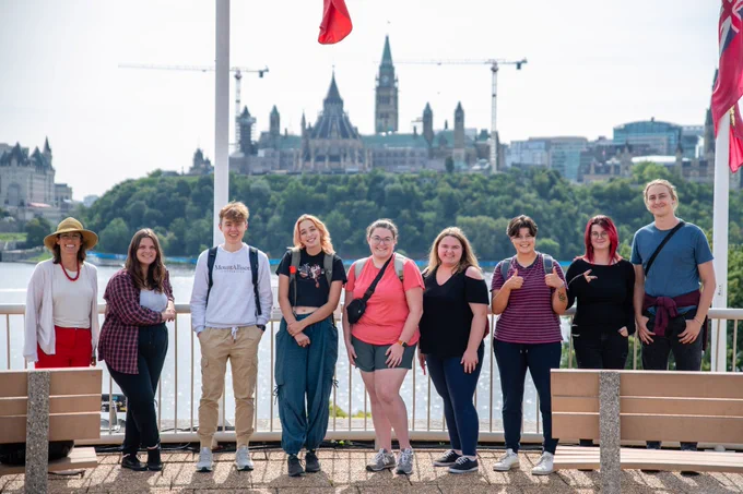 Outdoor photo of Dr. Susie Andrews and eight university students smiling and standing in a row on a pathway by the Ottawa River. In the distance is a treed riverbank, the Canadian parliament buildings, two construction cranes and an overcast sky.