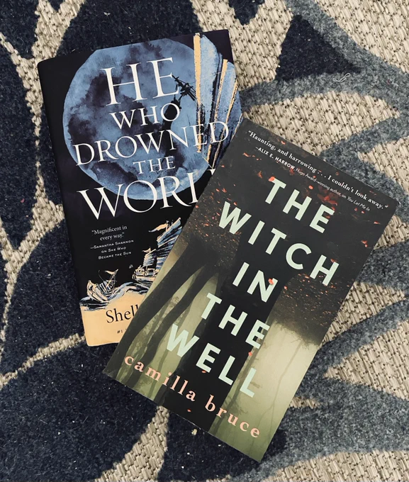 He Who Drowned the World and The Witch in the Well, stacked on top of each other on a blue and cream carpet