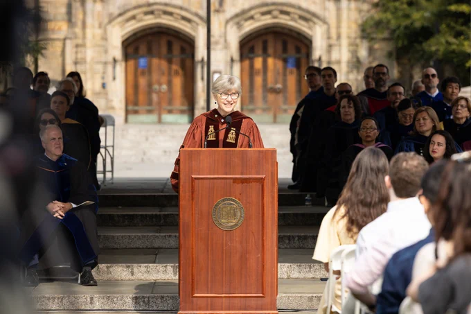 Photo of Lynn Cooley, dean of the Yale Graduate School of Arts and Sciences, stands behind a brown, wooden podium at the 2023 matriculation ceremony. Audience members in foreground. Other Yale dignitaries and Sterling Memorial Library in background.