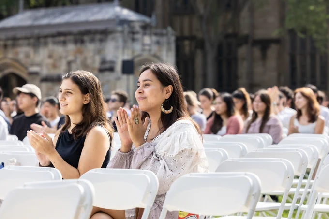 Photo of audience members sitting in white chairs at the 2023 Yale Graduate School of Arts matriculation ceremony. Stone buildings, green trees background.
