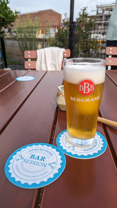 Ein Glas Bier steht auf einem Tisch im Freien, im Hintergrund die Speichergebäude des Dortmunder Hafens