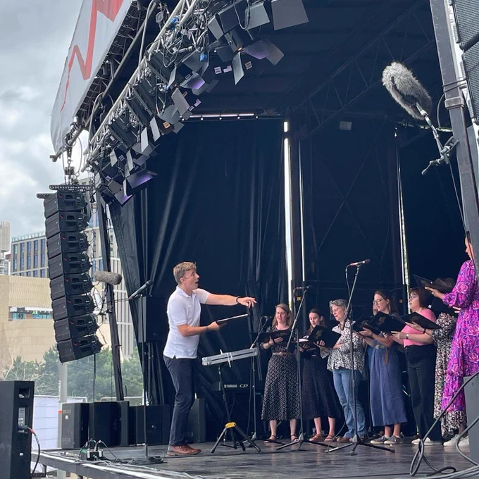 A side view of Ed, gesturing towards the BBC Singers as they sing. You can see the sopranos in the background, they are holding black folders containing sheet music.
