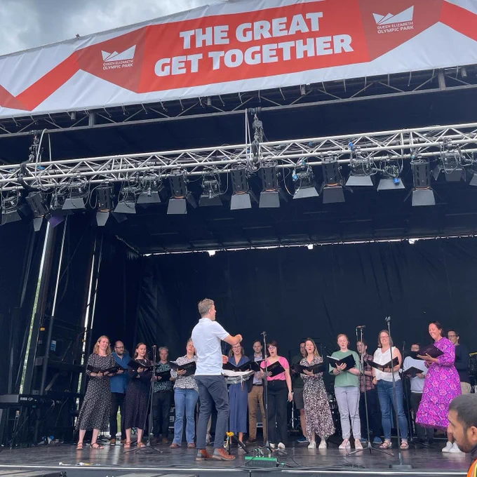 The BBC Singers on stage, led by Ed Price. The Singers are stood in a semi circle facing the audience, and Ed is facing the Singers.
