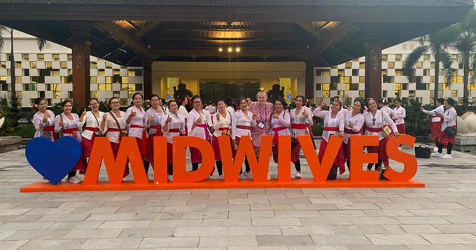 Midwifery student Meghan Critchlow poses with 15 women in traditional Balinese dress, behind red giant letters spelling out 'Midwives' at the  International Confederation of Midwives (ICM) Congress 2023 in Bali.