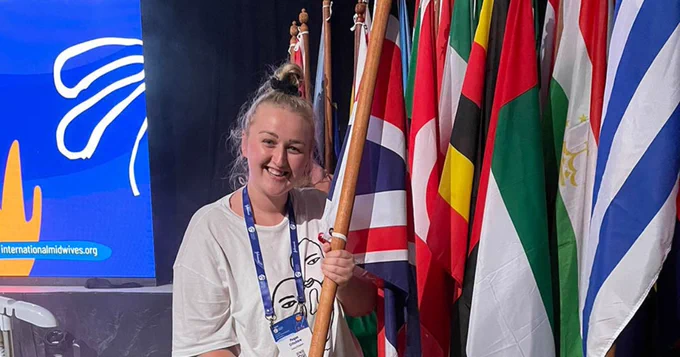 Midwifery student Meghan Critchlow smiles while holding a Union Jack flag, in front of several other national flags, at the International Confederation of Midwives (ICM) Congress 2023 in Bali.