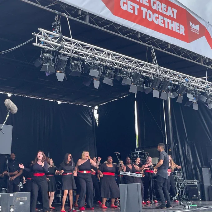 Stratford East Singers on stage. A banner above the stage reads 'The Great Get Together'. All the singers are wearing black with red accessories.