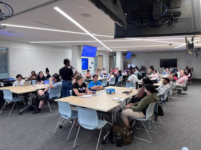 A group of about 55 individuals seated at tables in a training room listening to a presenter.