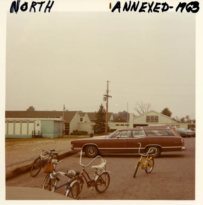 Color photograph showing the exterior of a one-story elementary school. A brown station wagon and four children's bicycles are parked in the foreground. Text written on top of the photographs reads "North Annexed - 1963"