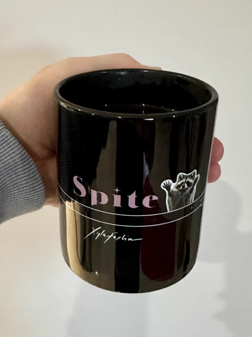 Photo of a black mug filled with black coffee. On the outside of the mug is text “Spite” in pink along with a rocket and raccoon.