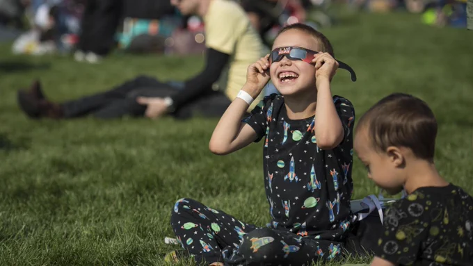 A young child sits in the grass and looks at the sky while wearing space-themed shirt and pants and holding eclipse-viewing glasses to his face.