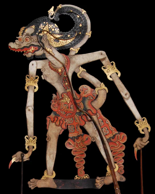 A colourfully painted two-dimensional shadow puppet of the character 'Trigangga', depicted in cream, red, gold and black. Thin bamboo poles are connected to his waist and wrist to allow the puppeteer to articulate it.