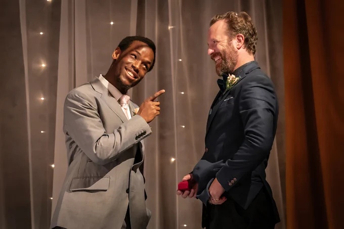 One actor holds a red velvet ring box as another actor points at him. They smile at each other.