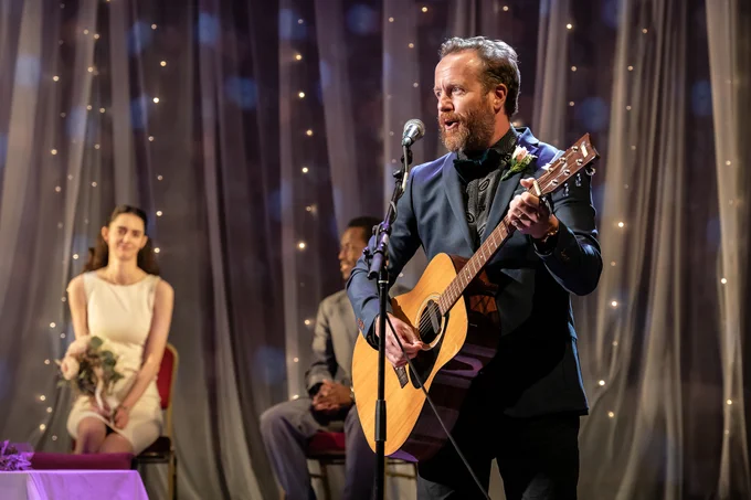 An actor plays the guitar and sings during a wedding scene.