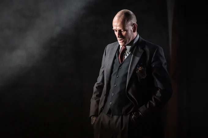 An actor wears a suit and looks down. Dramatic lighting.