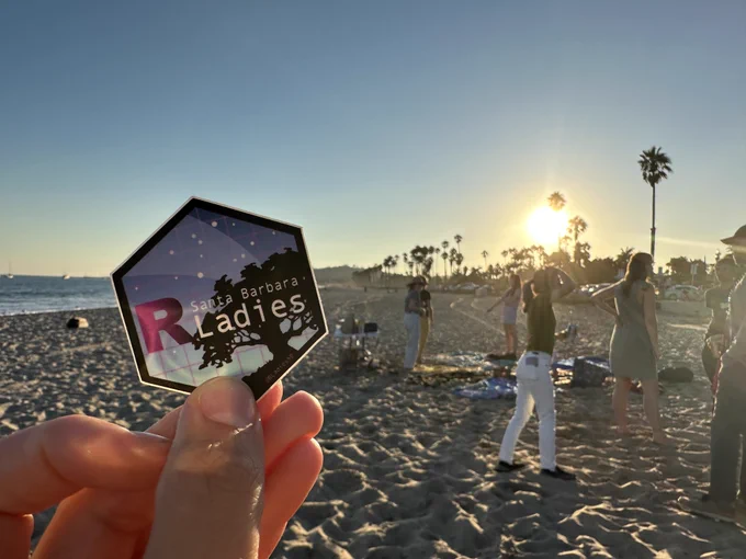 A hand holds up an R-Ladies Santa Barbara hex sticker. In the background, R-Ladies participants are scattered about the beach, conversing . Palm trees, the ocean, and the setting sun are the backdrop.