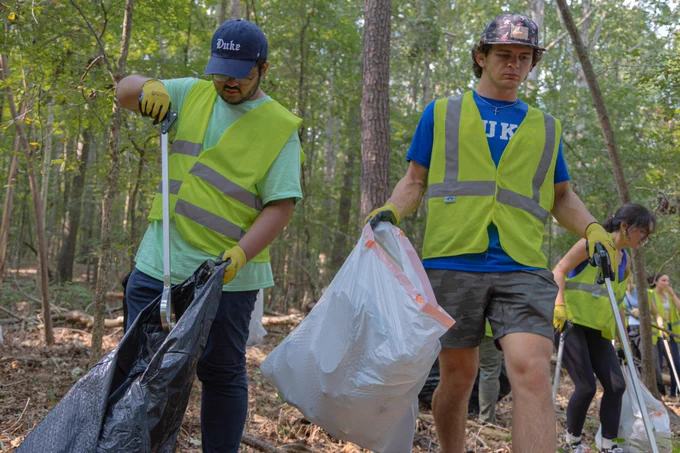 Two students in Duke gear and bright yellow vests pick up trash in the forest