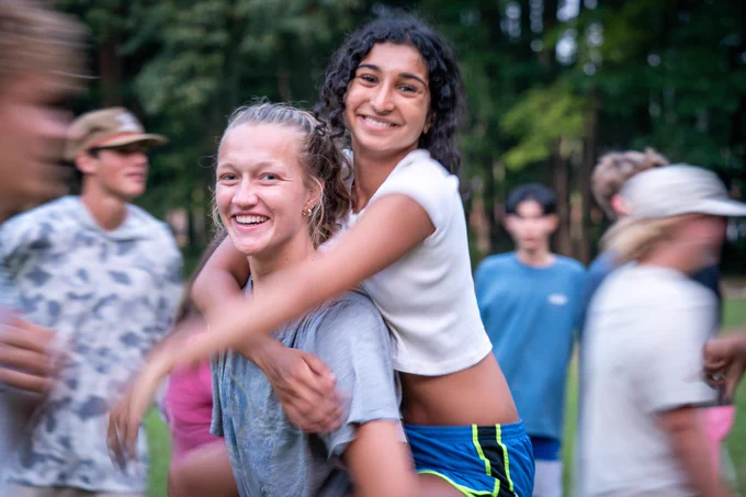 One student holds another on her back as they smile at the camera. There are other students around, blurred with movement.