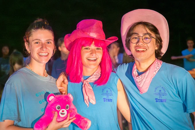 Three students in blue Project Waves Staff shirts pose for a photo. They all have bright pink accessories including a stuffed animal, wig and cowboy hats. 