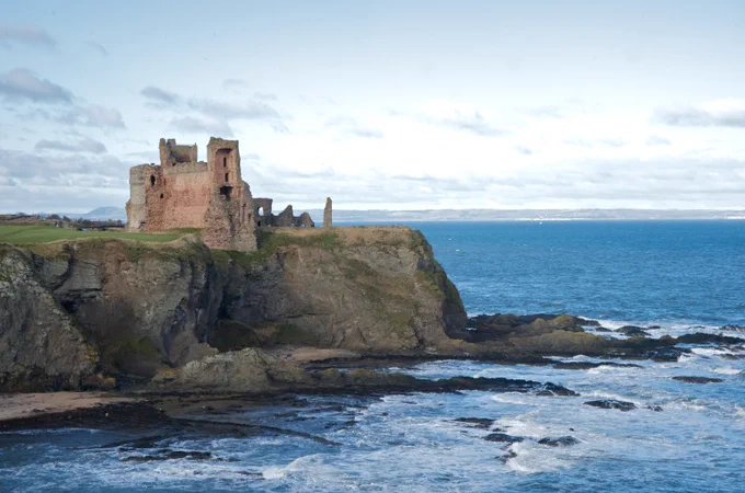 Image shows Tantallon Castle in East Lothian. The semi-ruied grey castle is perched on the cliff edge overlooking the sea. Below it, waves are breaking on rocks, and the cliff edge looks as if it is eroding into the water.
