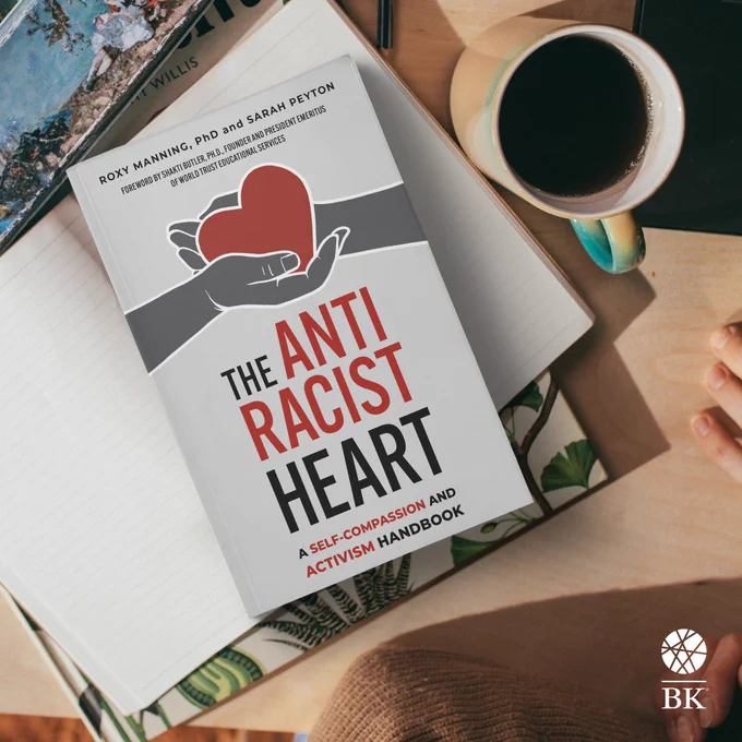 A copy of The Antiracist Heart sits on a desk atop an open notebook, with a cup of coffee nearby.