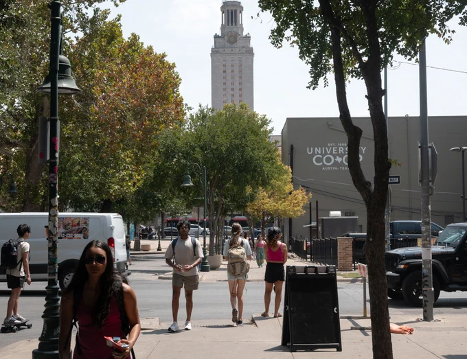 UT Austin students are walking along the sidewalk, some towards campus and some away from campus. These students are walking near the University Co-Op on a bright, sunny day with the Tower rising high above the campus. Photo by Leila Saidane.