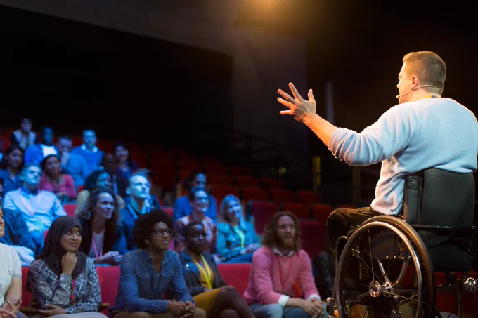 A man in a wheelchair on a stage is speaking to an audience. 