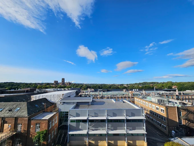 The view of Durham from the top of the Calman Learning Centre. A clear blue sky dotted with a handful of clouds acts as the background to Durham University buildings in the foreground. Durham Cathedral can be seen nestling amongst the treeline.