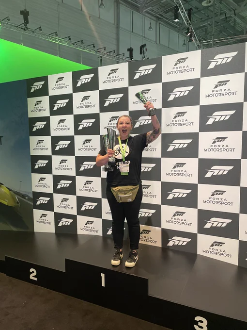 Kaitlyn standing on a the top of a winner’s podium from Forza Motorsport holding up multiple trophies raising her left arm in a victory pose.