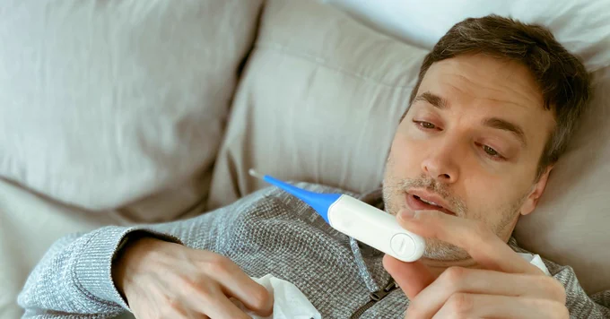 An image of a sick person in bed looking at a thermometer.