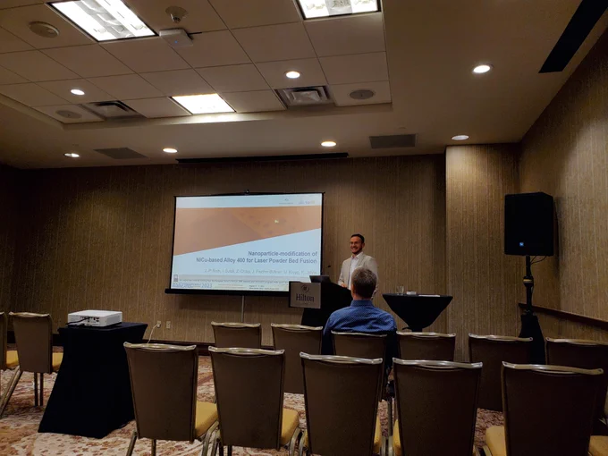 A young man stands at a podium and presents to a seated audience using a projector and powerpoint presentation on a screen.