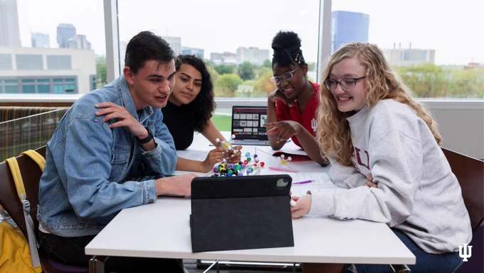 A group of students gather around a rectangular white table and look at an iPad.